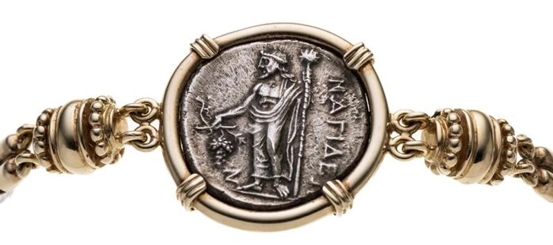 Zweireihiges Kordelarmband aus Silber im antikem Stil mit Gelbgold-Elementen. Mittig eine in Zarge gefasster Stater (VS: thronende Aphrodite, die eine Opferschale hält, hinter ihr steht Eros, RS: Dionysos stehend mit Stab und Trauben. Stadt Nagidos/Kilikien 374-356 v. Chr. ) . Armband ist beidseitig zu tragen. Durchmesser 2,7 cm. Vgl. Sear 5585.