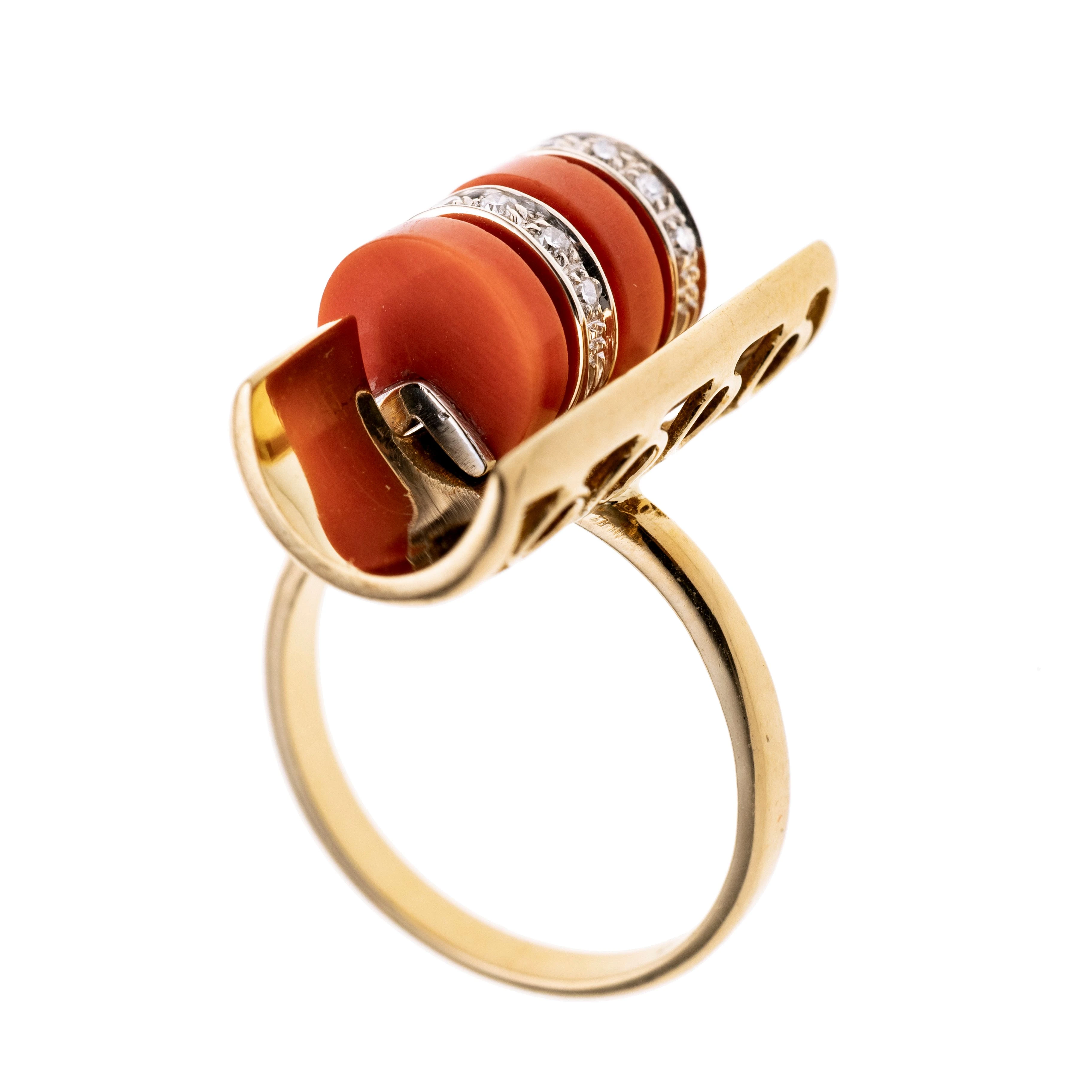 Schmale Ringschiene mit einem offenen halben rohrartigen Ringkopf vertikal angesetzt. Welcher seitlich mit Kreisen geschmückt ist und in dem Rohr sind Kreisscheiben aus Koralle mit dazwischen weißgold Elementen mit Diamanten besetzt.