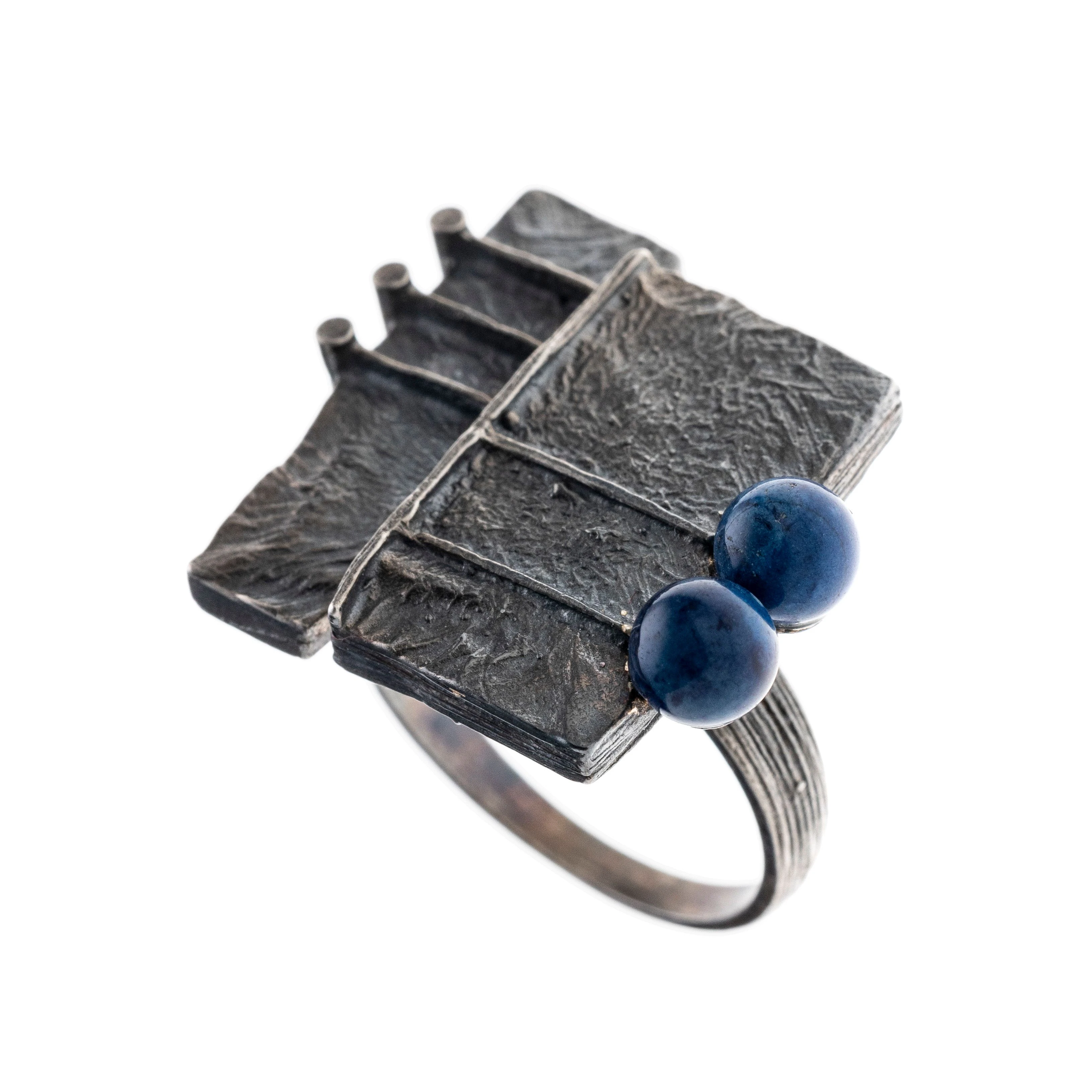 Ein silberner Ring mit quadratischen Elementen und unregelmäßiger Oberfläche als Ringkopf. Zwei Lapislazuliperlen sind als zusätzliche Zierde an einer Seite angeordnet.