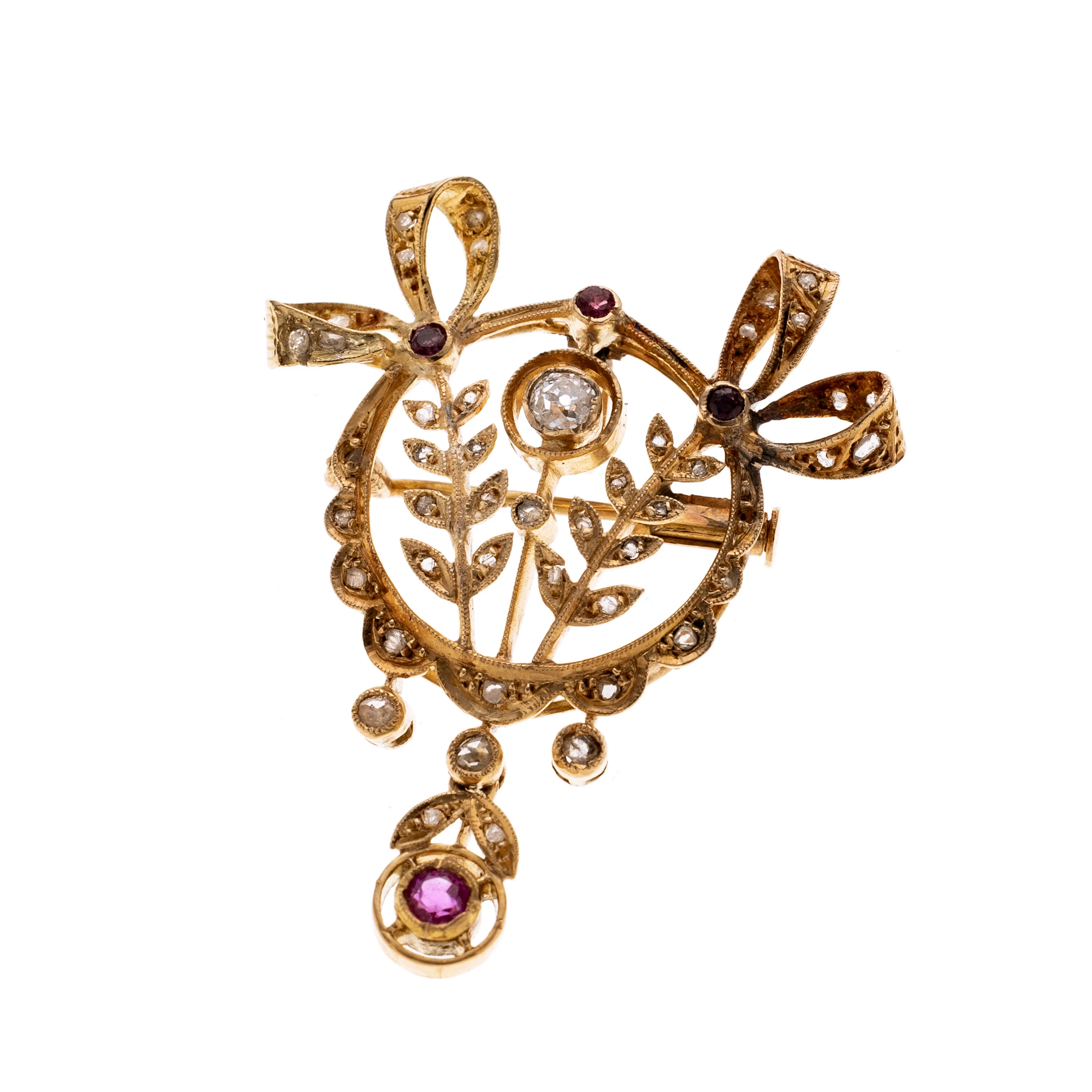 Ein durchbrochen gearbeiteter Kranz aus zwei Schleifenornamnenten ("Lovers Knot´s)", die mittig zweistilisierten Lorbeerzweige flankieren. Mittig befindet sich zu dem ein in Zarge gefasster Altschliffdiamant. Neben den Diamantrosen, die den Anhänger Zieren setzen drei Rubine und ein abgehängter Rubin einen Farbakzent. Der Anhänger ist in der klassisch-romantischen Formsprache gearbeitet unter der Verwendung der Symbolik für Beständigkeit, Sieg und Liebe. Länge mit Öse (moderner) 3,9 cm, Breite 3 cm.