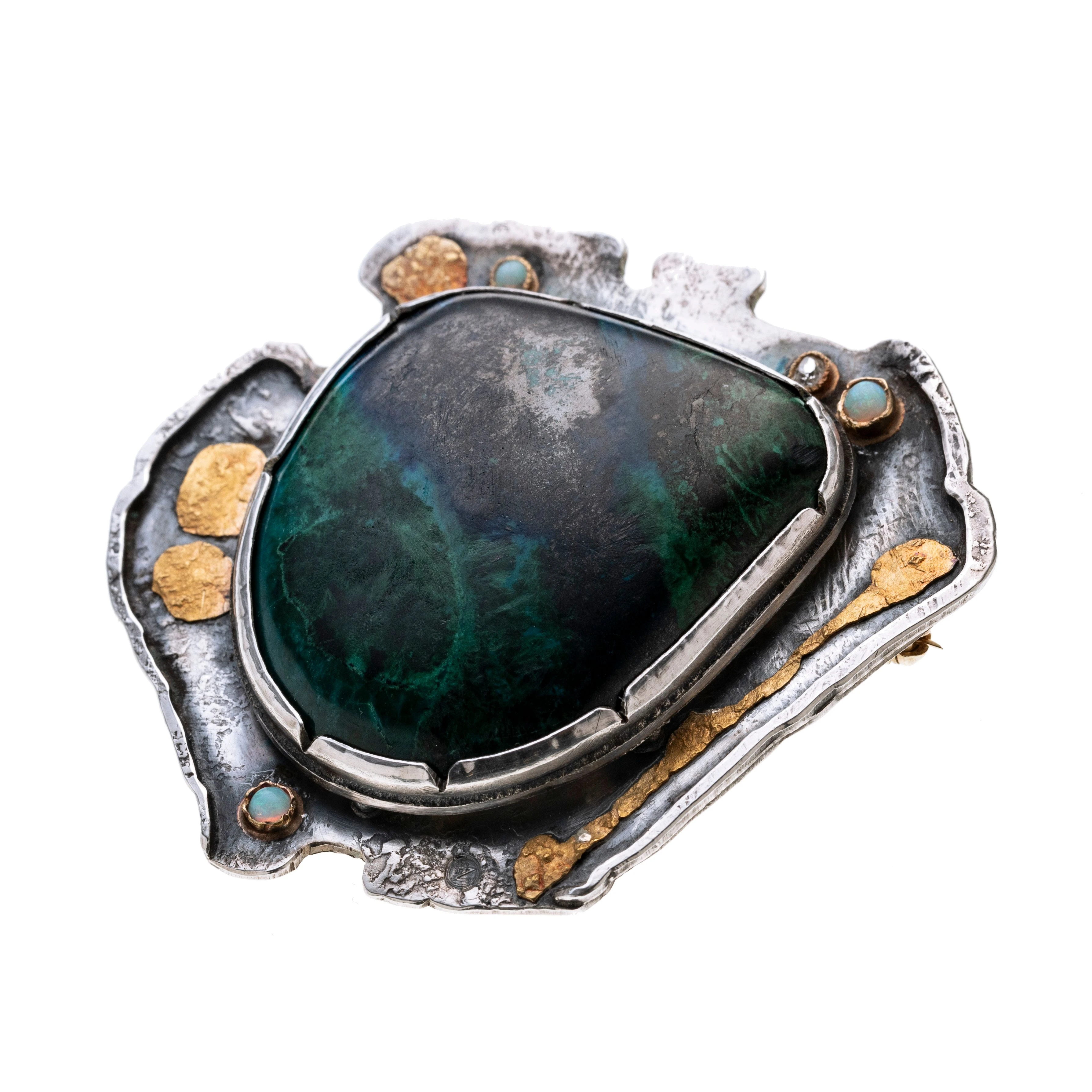 Ein Chrysokoll-Cabochon in in einer durchbrochenen Zargenfassung, mittig auf einer amorph gestalteten Form. Vier weitere goldene Zargenfassungen sind auf der Fläche aufgebracht, in denen drei Opale und ein Diamant gefasst worden sind. Goldene „Spritzer“ sind zusätzlich aufgelötet.